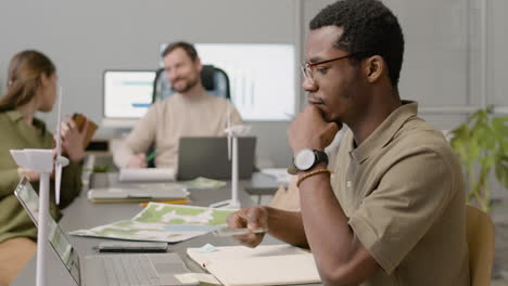 vista lateral de un hombre afroamericano que trabaja usando una computadora portátil y escribiendo notas sentado en un escritorio en la oficina. luego mira a unos colegas que están hablando de un proyecto