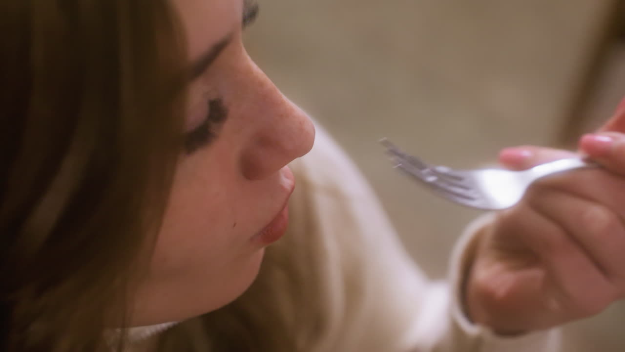 Close-up of girl placing creamy croissant topped with whipped cream and fruit into mouth with fork in cozy cafe. Enjoying delicious dessert with soft, warm atmosphere in background