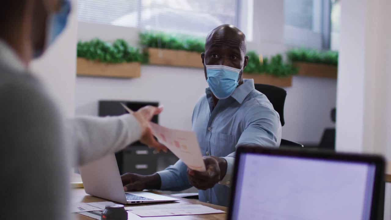 Diverse male and female business colleagues in face masks using laptops passing document in office