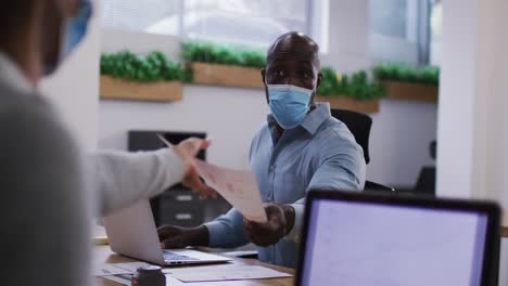 Diverse-male-and-female-business-colleagues-in-face-masks-using-laptops-passing-document-in-office