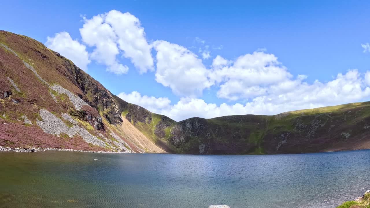 Clouds drift across a bright blue sky above Loch Brandy, with sunlight shifting over hillsides in a wide, steady landscape time-lapse shot
