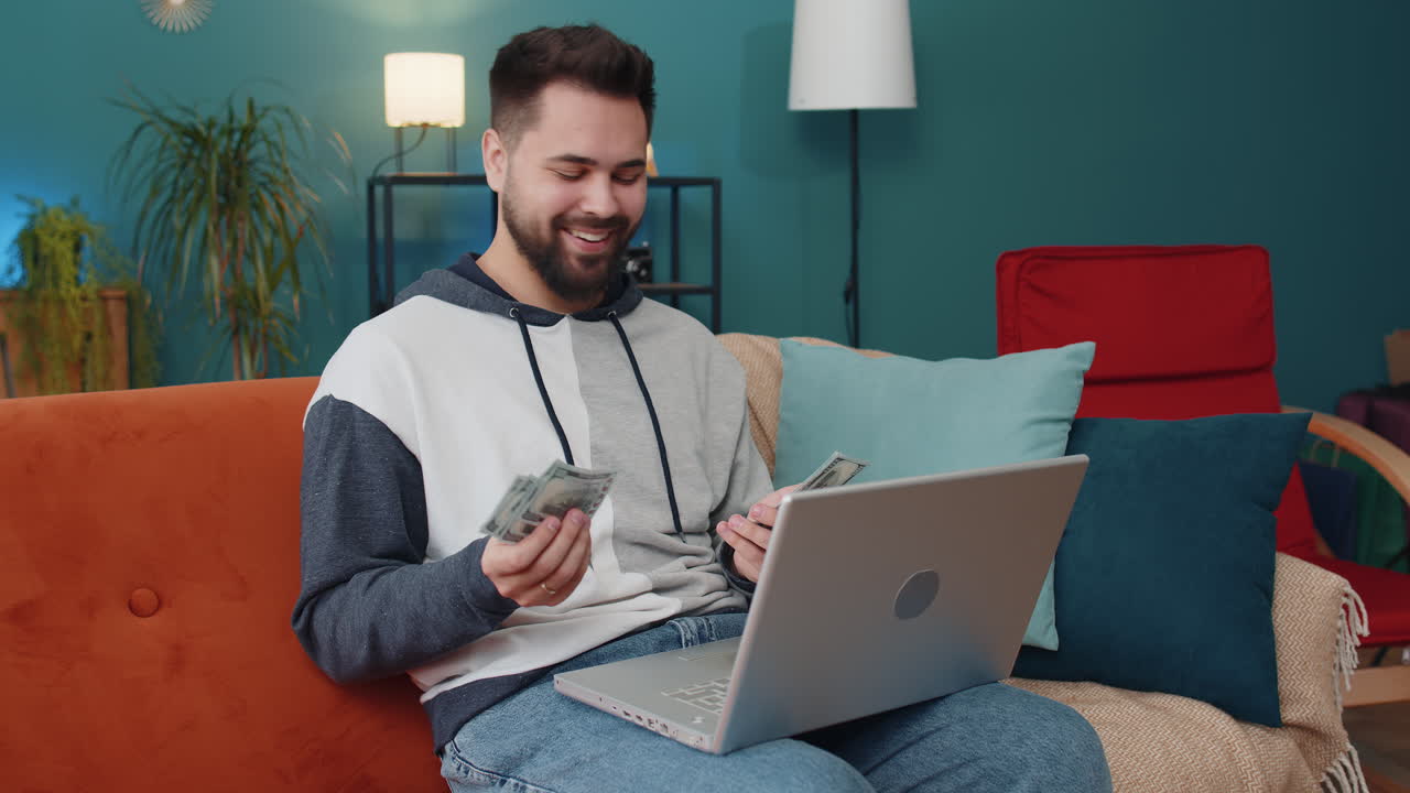 Smiling young man counting money cash and use laptop pc calculate domestic income earnings at home