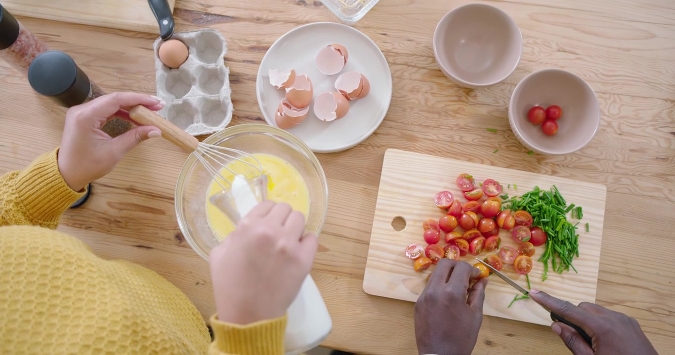Coppia afroamericana che prepara la colazione a tavola, uno sbatte le uova gialle, l'altro taglia i pomodori verdi.