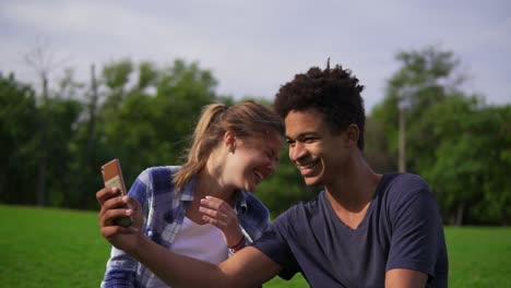 cute friends resting in a park, sitting on the grass, looking at the smartphone and laughing. mixed race couple sitting on the