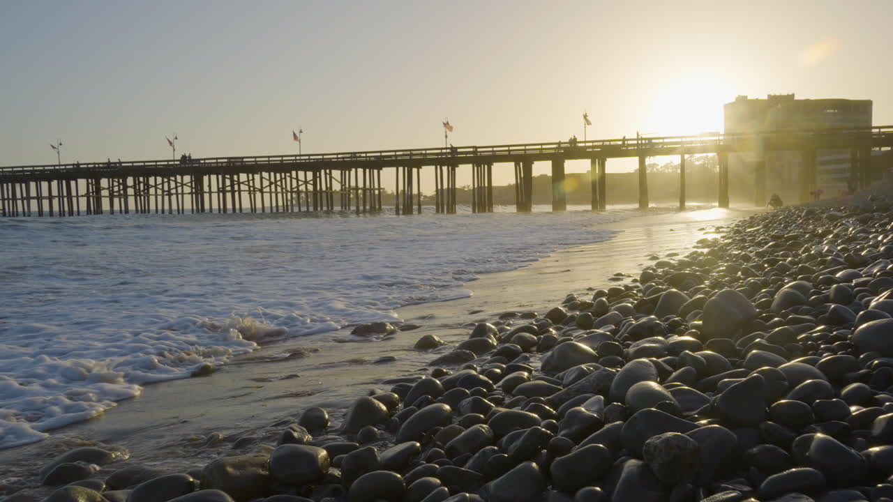 Carrellata in avanti lungo la spiaggia di Ventura mentre le onde si infrangono sulla riva con il molo di Ventura sullo sfondo, situata nella California meridionale