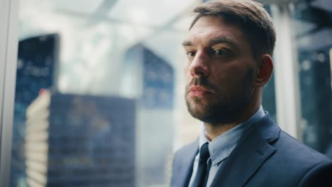 portrait of successful businessman in a suit riding glass elevator to office in modern business center. young male looking at modern downtown skyscrapers out of the panorama window in the lift.