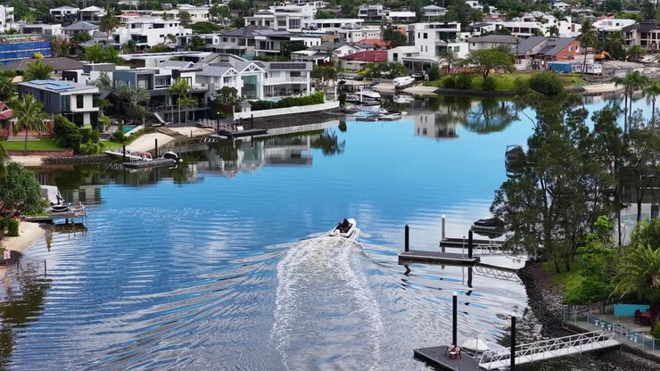 A boat navigates tranquil Gold Coast canals, reflecting luxury homes and clear skies, captured with smooth aerial motion
