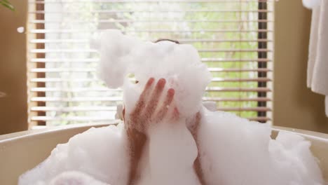 portrait of happy biracial woman lying in bathtube, blowing foam and pampering herself