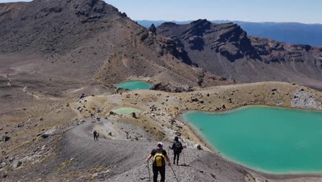 tourists hike down loose volcanic pumice trail toward emerald lakes