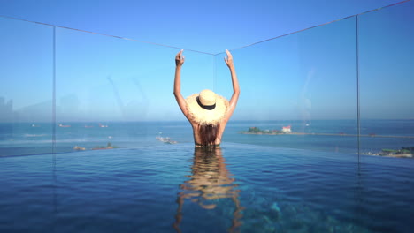 Back-view-of-a-slim-woman-standing-in-a-swimming-pool,-wearing-a-straw-hat-and-raising-her-arms-in-the-air,-looknig-at-infinite-ocean