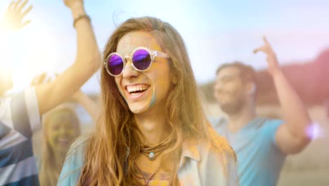 close-up portrait of a beautiful young girl with sunglasses standing in the crowd of people celebrating holi festival. people throwing colorful powder in her back. her face and clothes are covered with colorful powder.