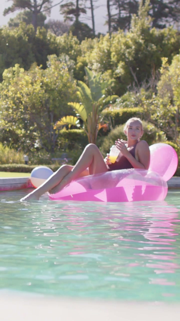 Vertical video: Sunning woman orange suit lying on pink float at pool sipping cup moving looking