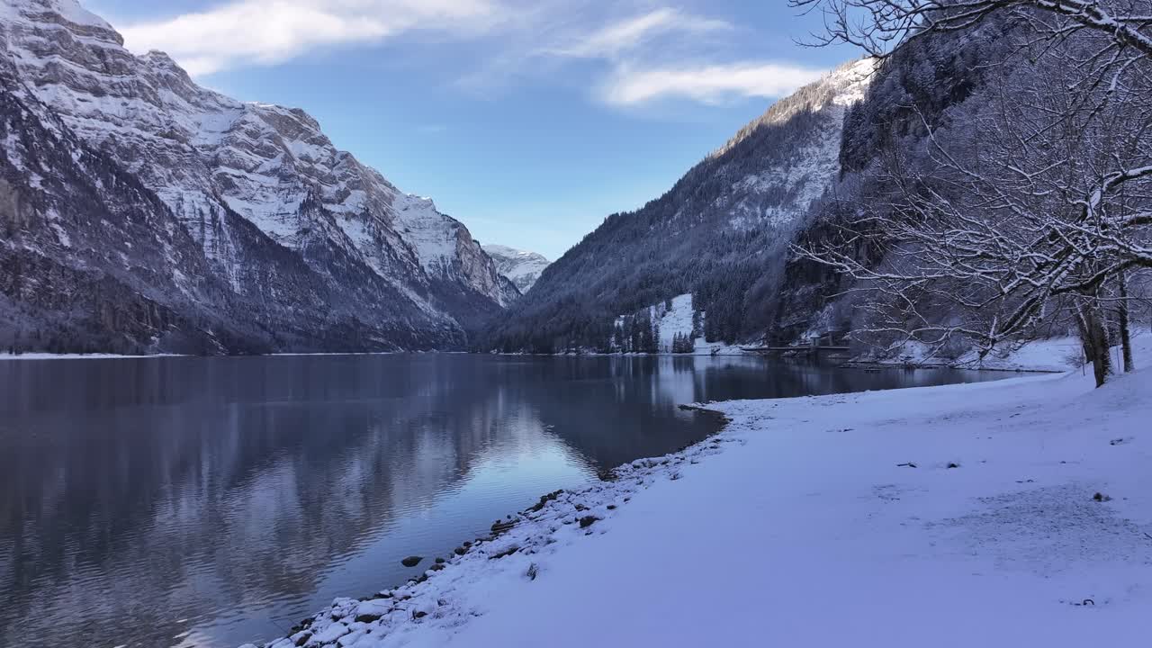 Drone pushes towards to tranquil Klöntalersee lake surrounded by majestic snowy Vorderglärnisch mountain in Glarus, Switzerland.