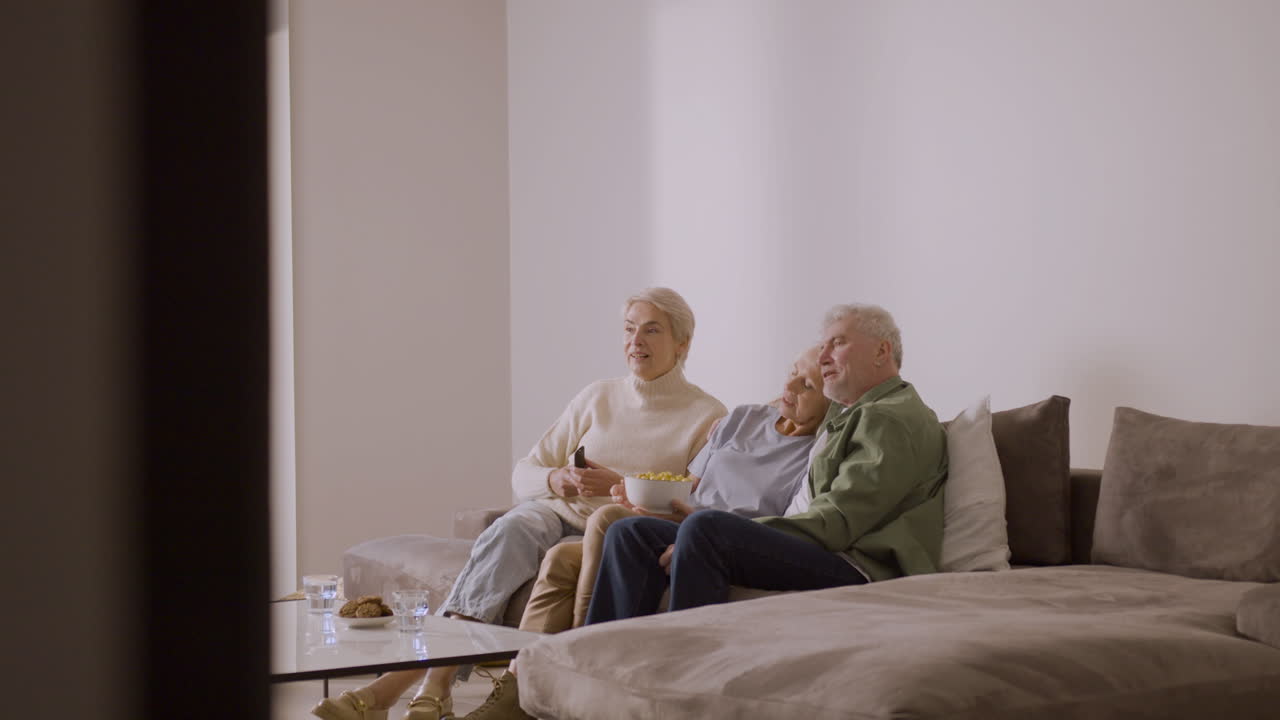 Trois personnes âgées regardant la télévision, discutant et mangeant du pop-corn au caramel, assises sur un canapé à la maison.
