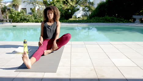 happy biracial woman practicing yoga sitting by pool in sunny garden, copy space, slow motion