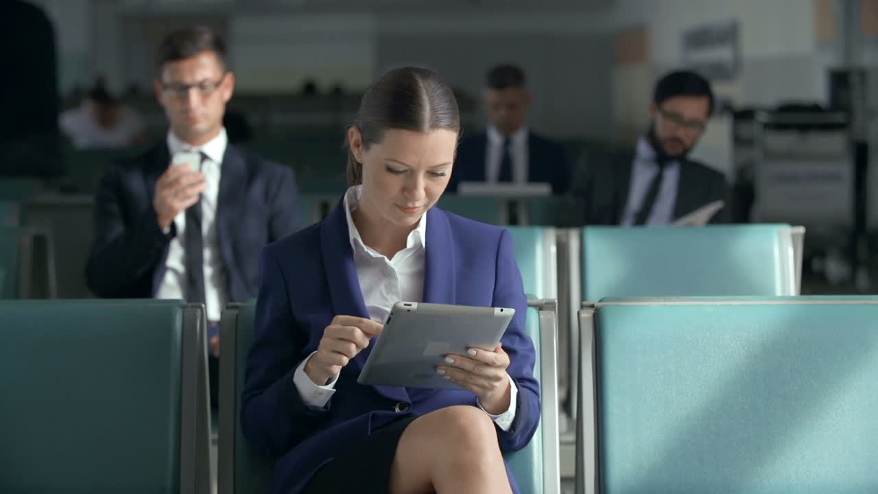 Business Woman In Suit Using A Tablet While Sitting In The Airport Waiting Area, Behind Are Several Men In Suits Sitting
