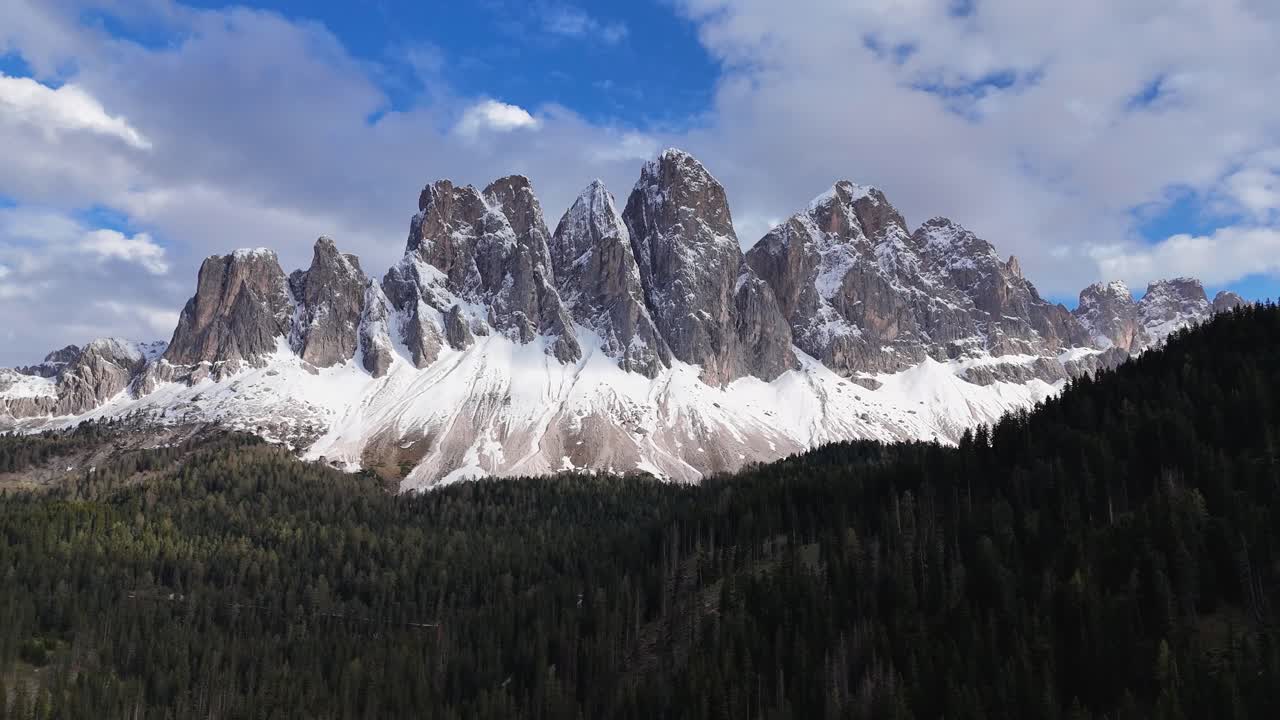 Odle Geisler Group in the Dolomites displays jagged peaks with snow, surrounded by a dense alpine forest under a partly cloudy sky in South Tyrol