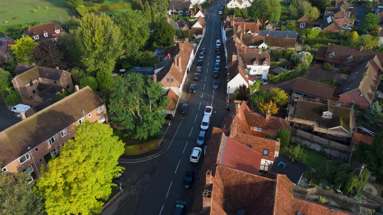 Drone flight over Dorchester on Thames village. The camera follows the curved road, showing charming old rooftops, cars, and leafy trees framing this picturesque Oxfordshire English countryside scene