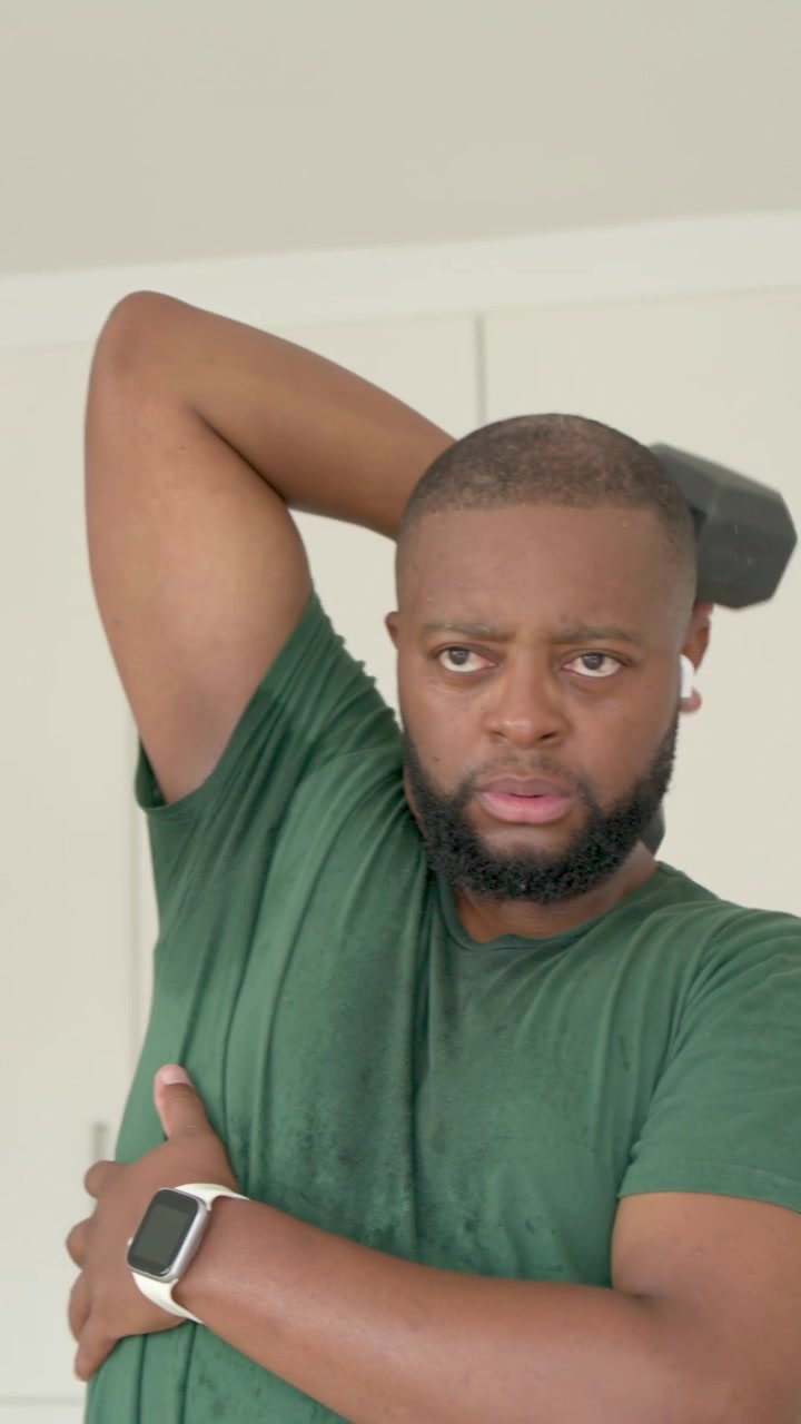 Vertical video: Lifting hex dumbbell, man in green tee exercising triceps at home, camera widening
