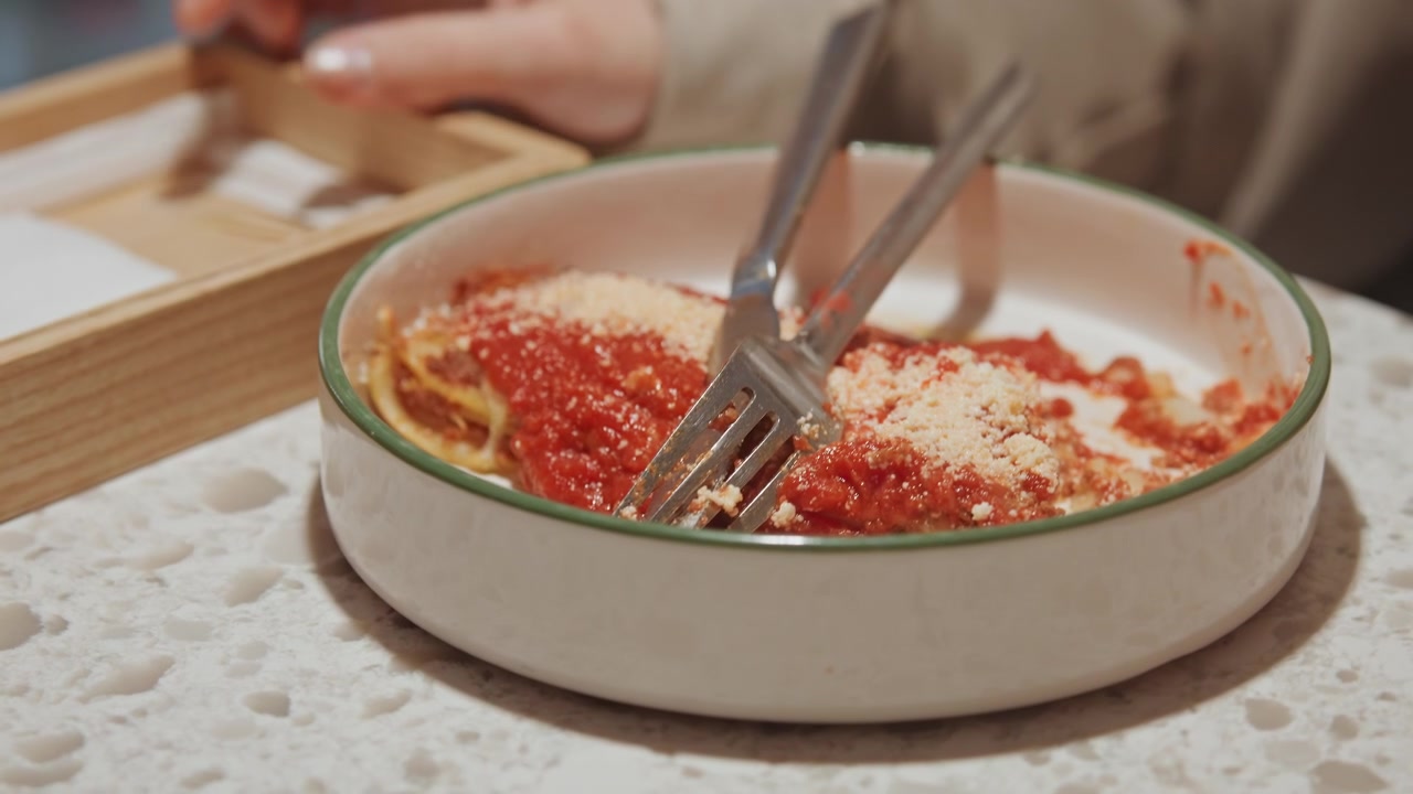 Primo piano di persona che usa forchetta e coltello per tagliare polpette coperte di salsa rossa su un piatto. Le mani interagiscono con gli utensili sul cibo, rivelando dettagli di texture come il formaggio grattugiato in superficie.