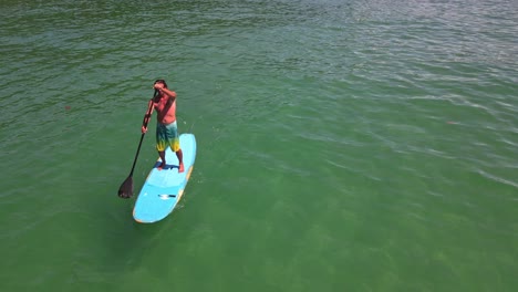 toma aérea estática de un hombre caucásico haciendo ejercicio en una tabla de remo se acerca a la cámara en aguas turquesas tropicales claras