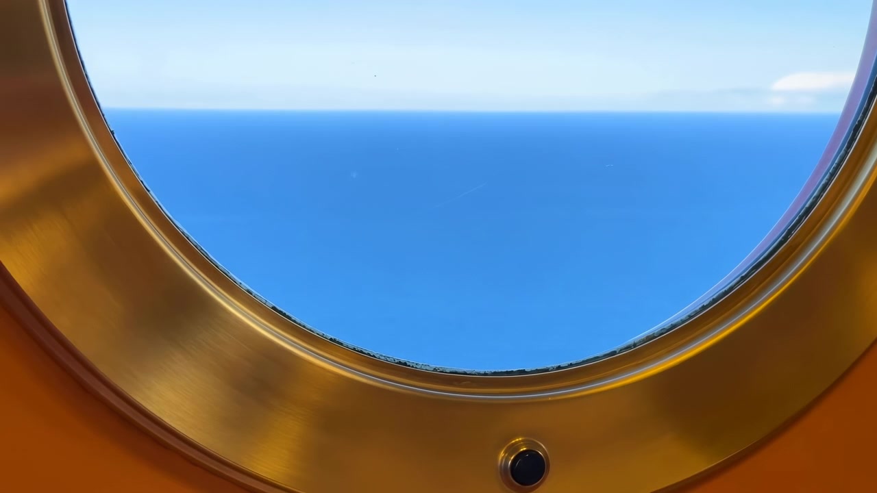 Primo piano centrato attraverso un oblò rotondo rivela un'ampia fascia di mare blu calmo e cielo pallido, con bordo in ottone spazzolato, toni caldi della cabina, luce diurna soffusa e un'atmosfera di viaggio tranquilla, con oblò e oceano.