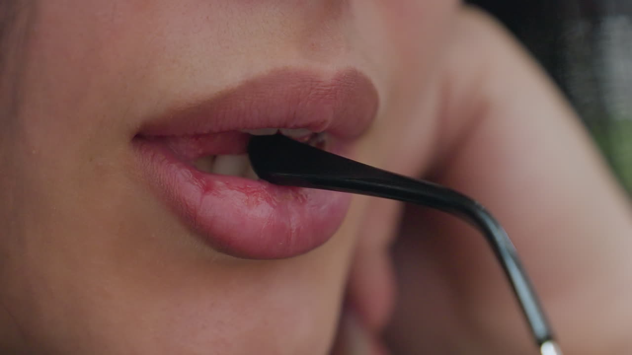 Close up of young lady gently biting eyeglass arm with soft smile on lips, showing relaxed facial expression and sensual detail in warm lighting with shallow depth of field