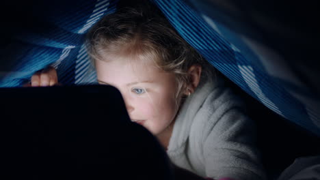 happy little girl using digital tablet computer under blanket enjoying learning on touchscreen technology playing games having fun at bedtime