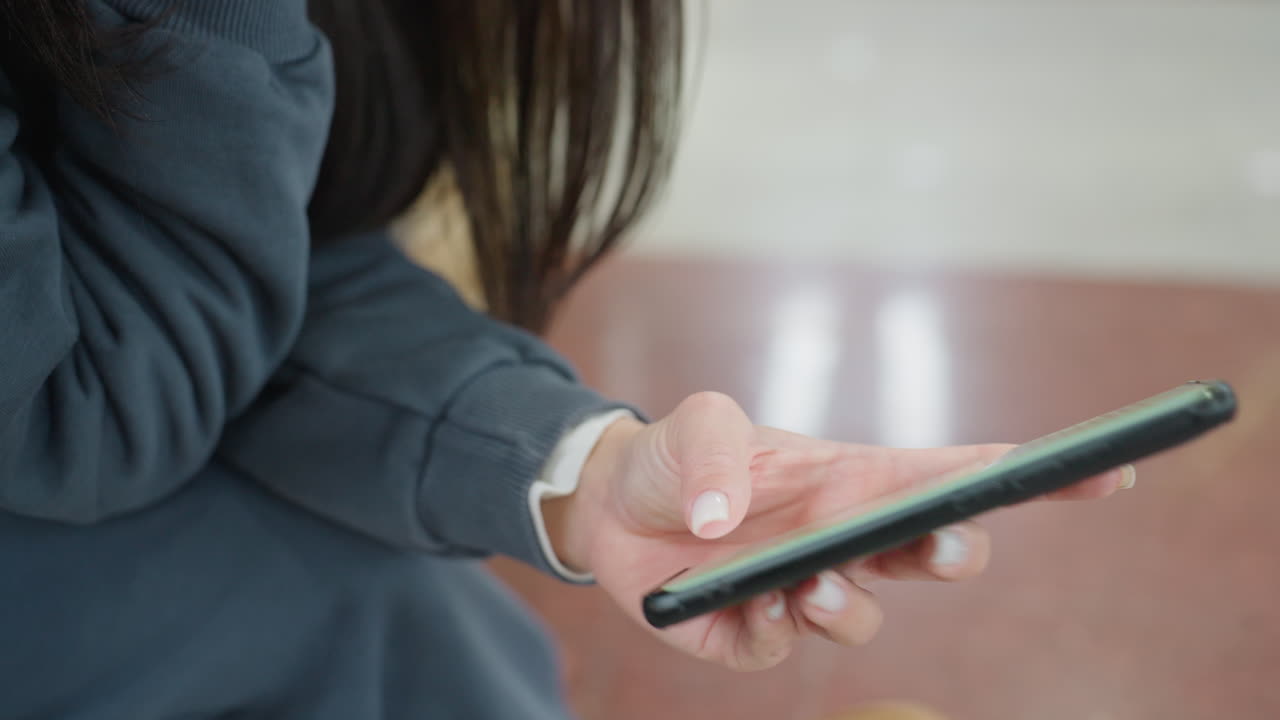 Close up of lady in dark casual outfit sitting and operating smartphone using both hands, focus on fingers and phone screen, soft indoor light with bokeh plant and reflective floor in background