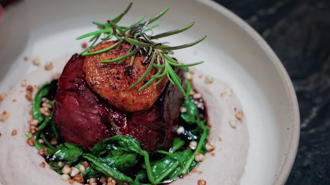 Close up of a gourmet dish featuring grilled steak topped with foie gras, served with sauteed spinach and a rosemary garnish