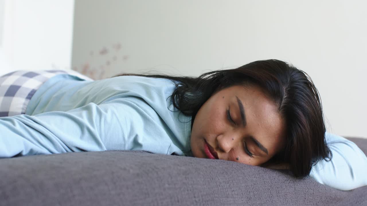 Donna asiatica di mezza età che si sveglia, solleva uno smartphone bianco per controllare e si riaddormenta su un divano grigio