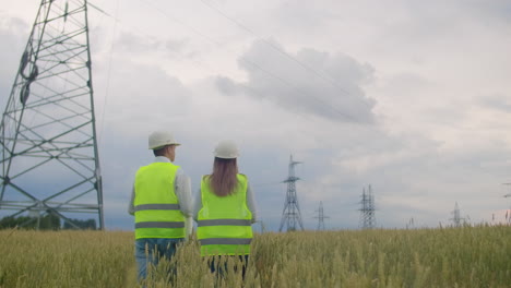 The-view-from-the-back:-group-of-engineers-at-a-high-voltage-power-plant-with-a-tablet-and-drawings-walk-and-discuss-a-plan-for-the-supply-of-electricity-to-the-city.-Transportation-of-renewable-clean-energy.