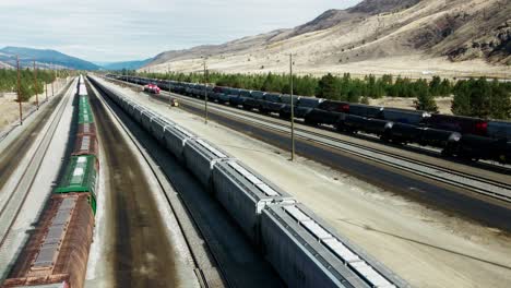 sideways-flight-dolly-drone-shot-flying-between-cargo-trains-and-tank-trains-on-a-railroad-station-in-a-desert-environment-on-a-sunny-day-with-mountains-in-the-background-and-powerlines