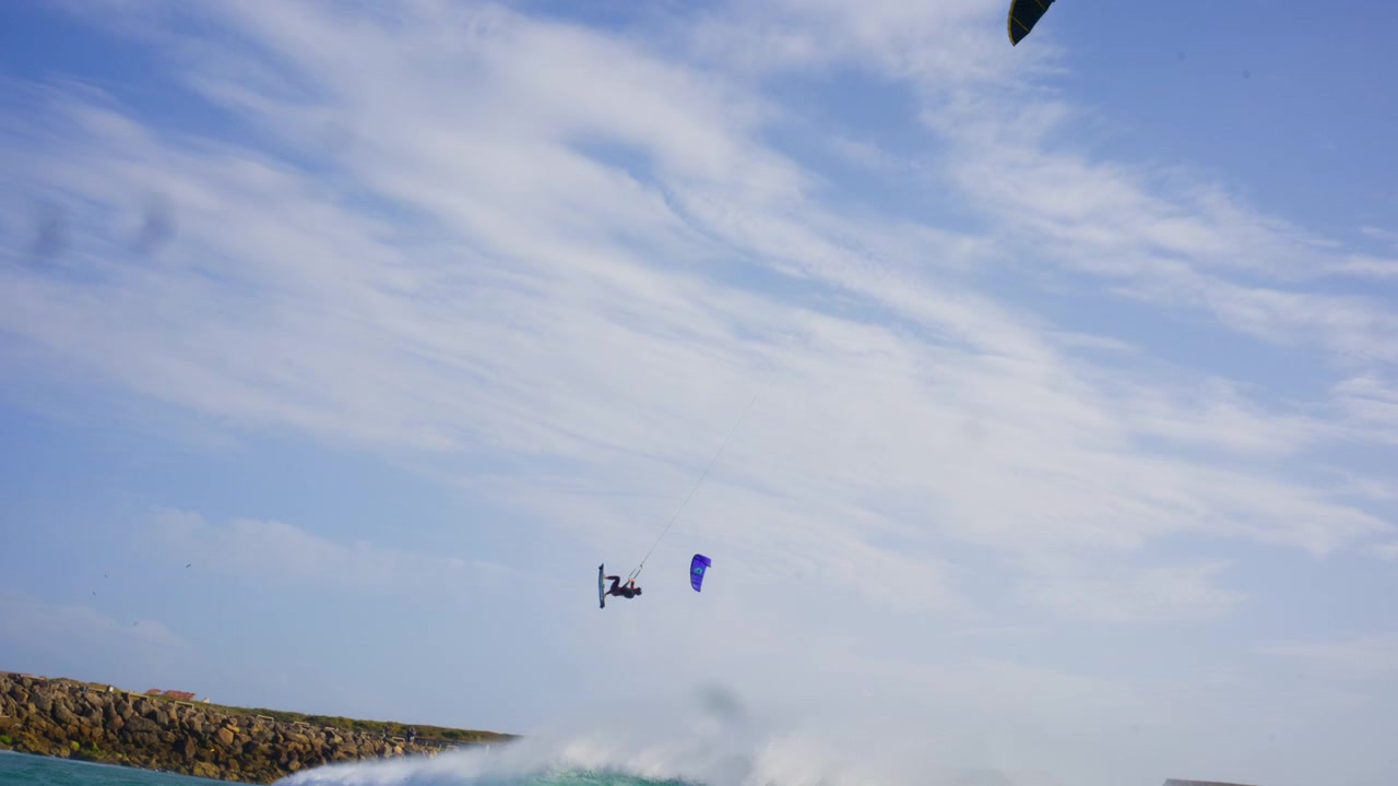 Bred optagelse i vandoverfladehøjde af Balneario, Tarifa med Isla de Tarifa i baggrunden. Professionelle kitesurfere laver enorme big air-spring i den legendariske Levante-vind, mens bølgerne bryder langs kystlinjen