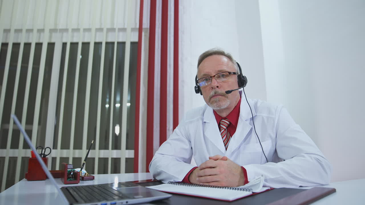 Older doctor listens to patient during video call. Senior male physician in medical uniform and headset having a remote consultation. Telemedicine concept.