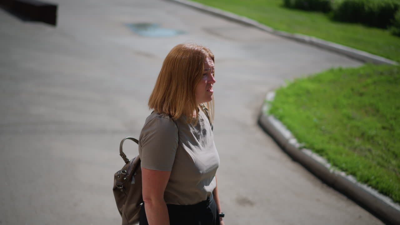 Aerial view of unhappy girl standing under bright sunlight near school building, looking around and checking time with fitness bracelet, showing sadness and contemplation in warm summer atmosphere