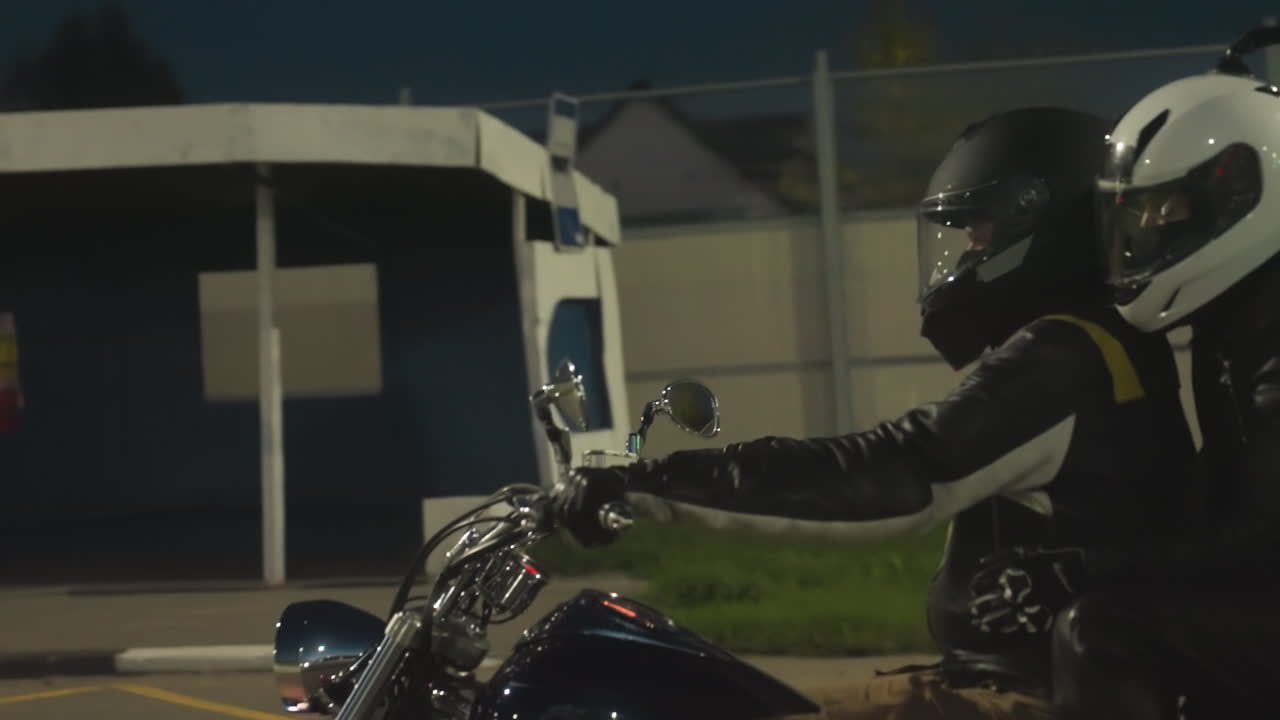 Back view of motorcycle rider wearing helmet and leather jacket crossing intersection under glowing green traffic light at night, passing pedestrian crossing with illuminated road signs