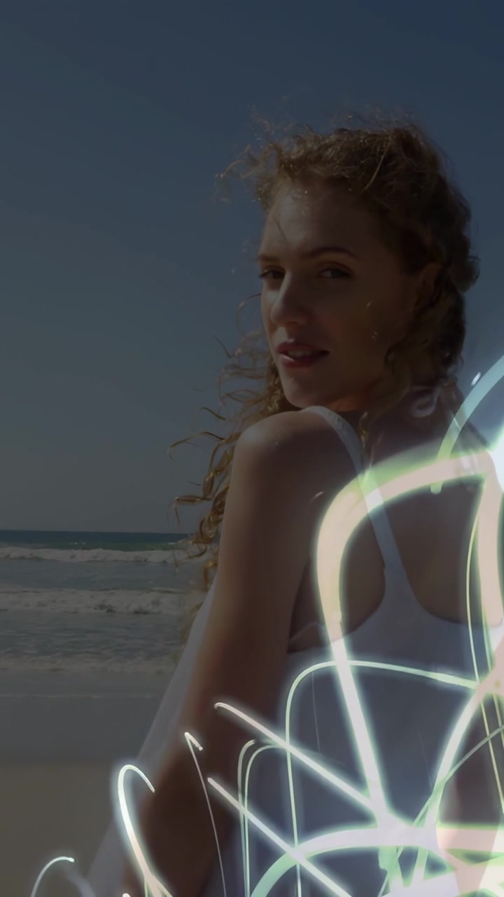 Vertical video: Woman turning to cam as neon trails rising on beach making turn showing dress ads