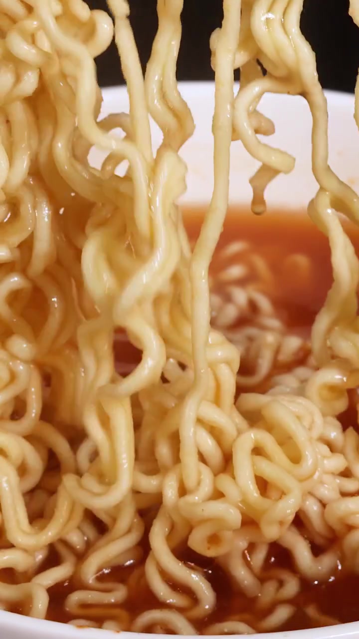 Le bacchette sollevano noodles istantanei ricci da una ciotola fumante di brodo, con primi piani sotto un'illuminazione brillante e uniforme e un'angolazione della telecamera statica.