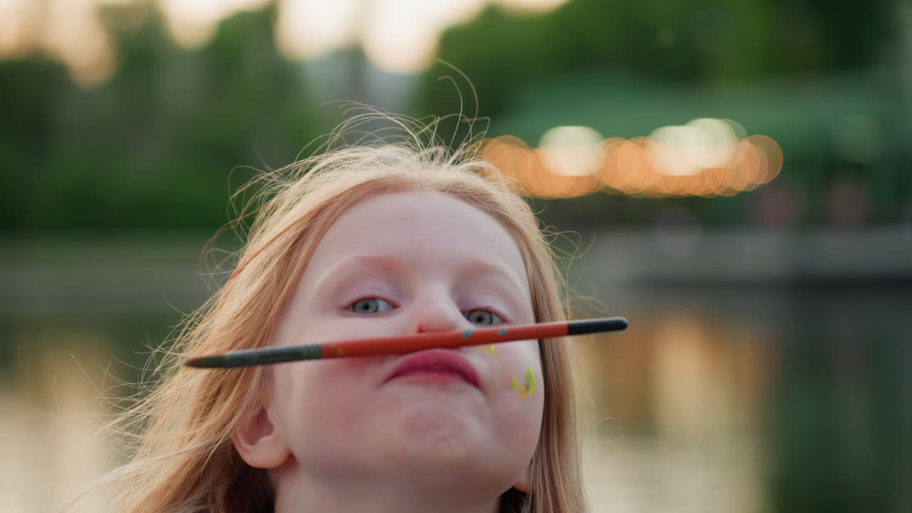 close up of child balancing pencil on nose with playful grin in summer park by lake, blurred adult behind, paint dots on cheek, warm twilight glow, fun creative moment of carefree family