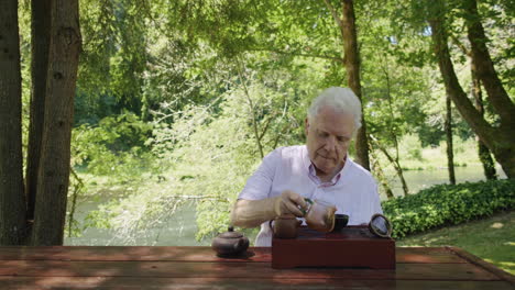 older man pours tea from pitcher, sips tea, looks toward river, teapot