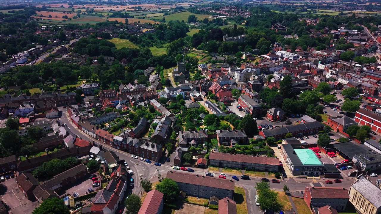 Hohe Luftaufnahme über Devizes in Wiltshire, die historische Gebäude, einen Kirchturm, Wohnstraßen, Gärten und grüne Ackerflächen zeigt, die sich an einem sonnigen Sommertag über die Marktstadt hinaus erstrecken