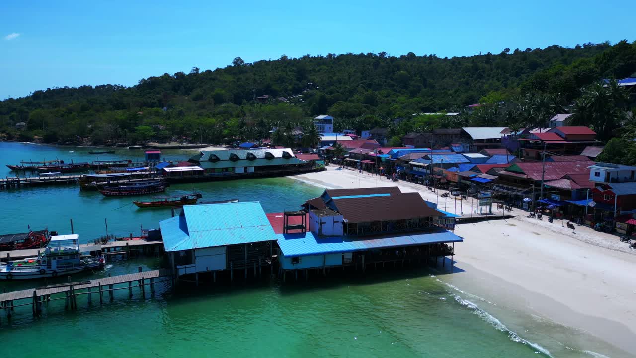 Palm trees projecting shadows on the white sand beach of Koh Rong Island village in Cambodia, with colorful houses and lush vegetation.Breathtaking aerial view flight static tripod hovering drone