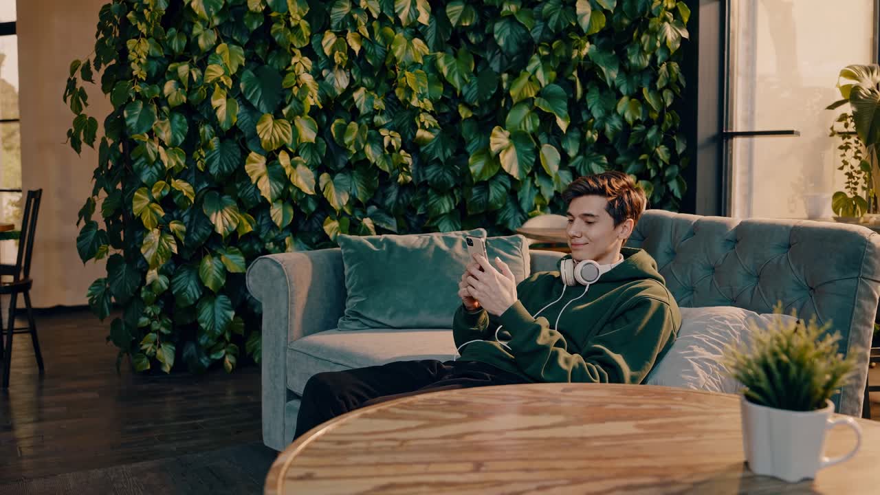 relajándose dentro de un café moderno, joven sentado cómodamente en el sofá cerca del jardín vertical, navegando por el teléfono inteligente con auriculares inalámbricos