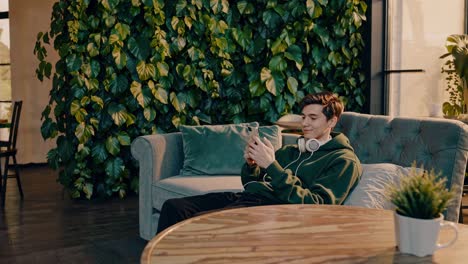 relaxing inside modern cafe, young man sitting comfortably on couch near vertical garden, browsing smartphone with wireless headphones