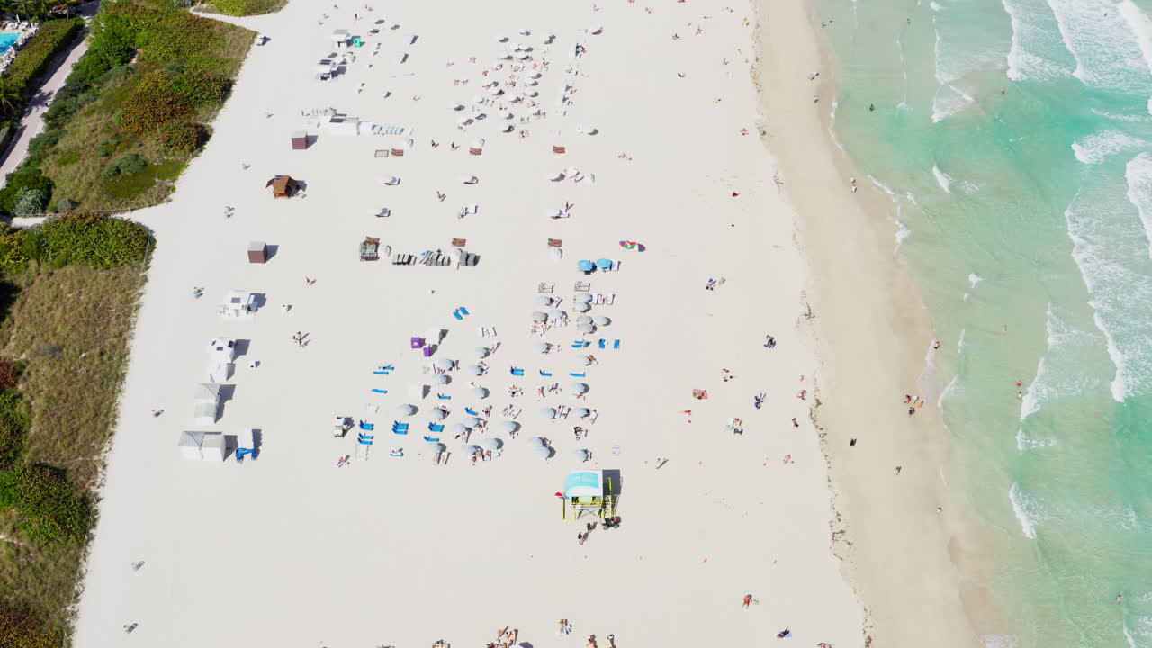 A top-down drone view vividly captures sunbathers, umbrellas, and waves on the iconic sands of South Beach, Miami.