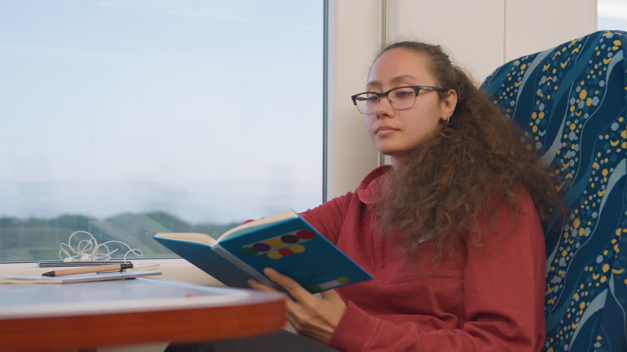 Jeune femme lisant près de la fenêtre d'un train, cheveux bouclés et pull rouge, livre de poche ouvert sur les genoux avec des lunettes sur la table, campagne défilant à l'extérieur, ambiance sereine et contemplative capturant le voyage lent et le confort.