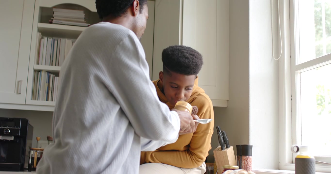 Padre afroamericano che preleva da un barattolo con un cucchiaio mentre il figlio adolescente assaggia la crema spalmabile al bancone della cucina.