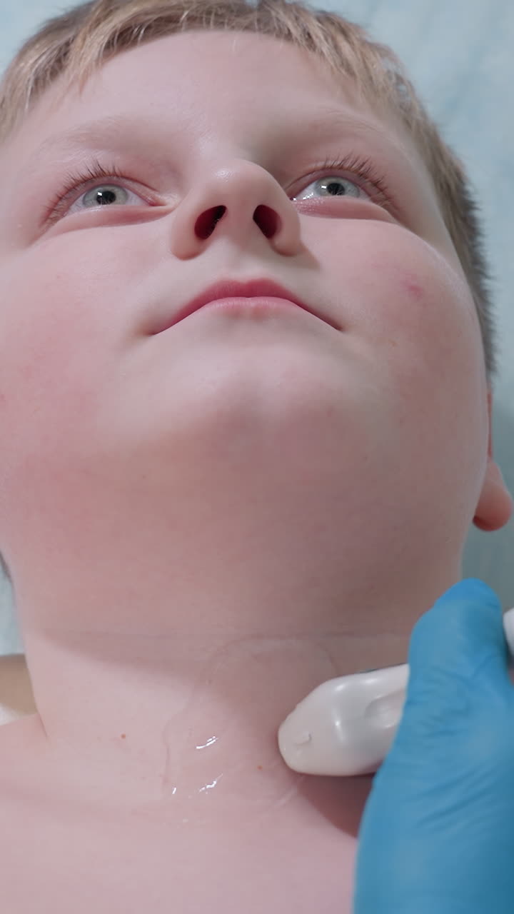Primer plano de un niño al que se le realiza una ecografía de cuello; el ecografista desliza la sonda sobre la tiroides con gel mientras mantiene las manos enguantadas firmes; imagen en tiempo real que se muestra en el monitor; evaluación centrada de las glándulas