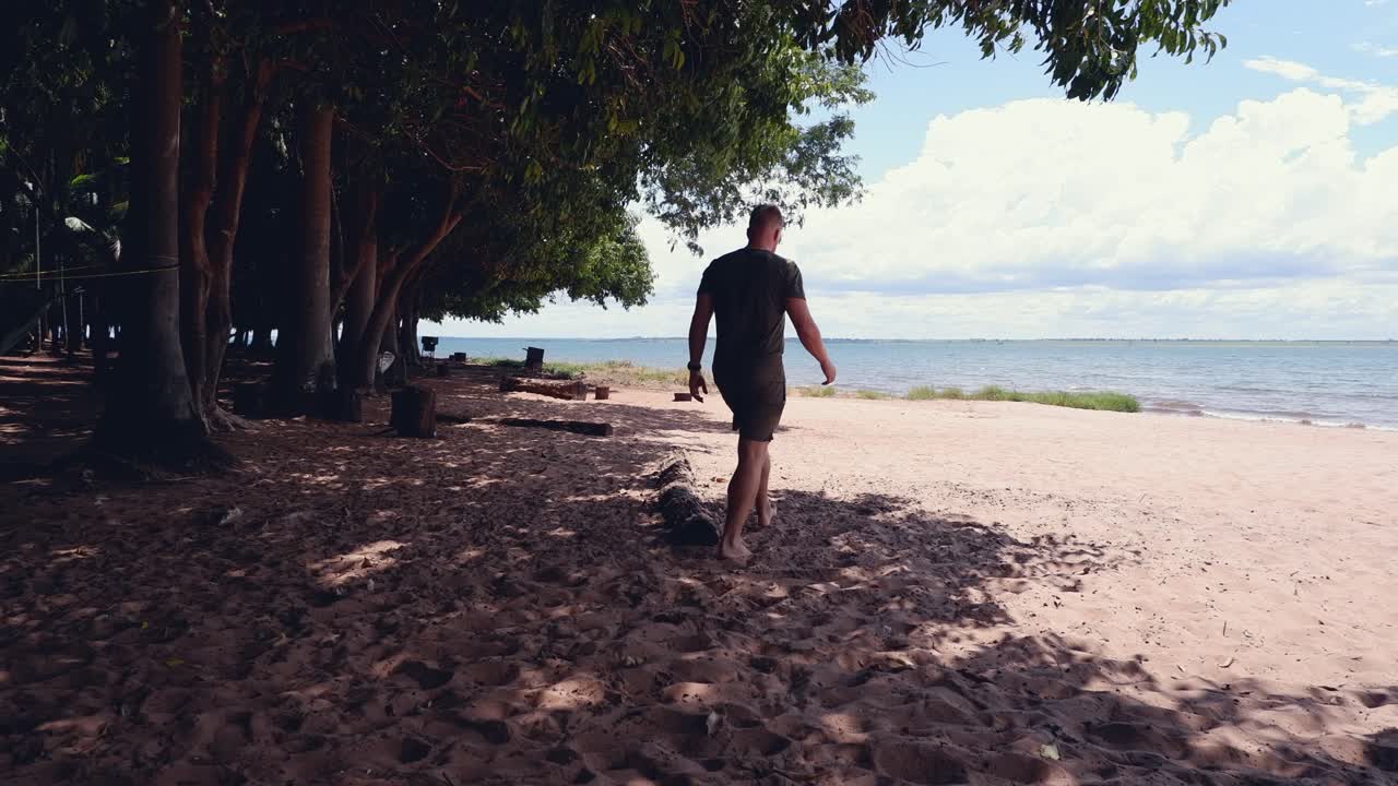 Caucasian man walks from shaded campsite onto sunny, sandy beach